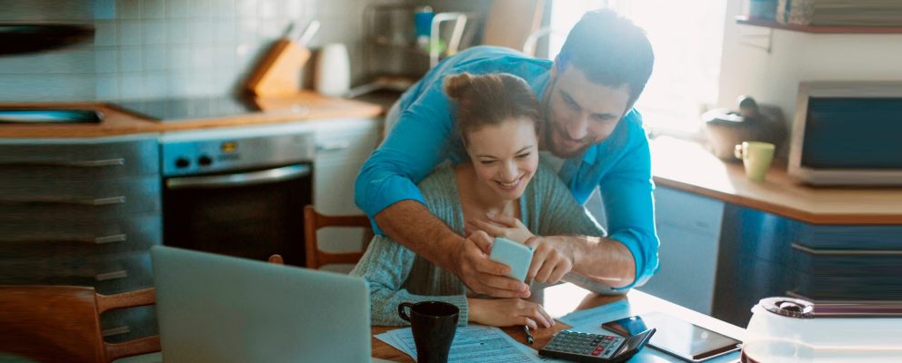Couple looking at a mobile phone while reviewing paperwork in their kitchen