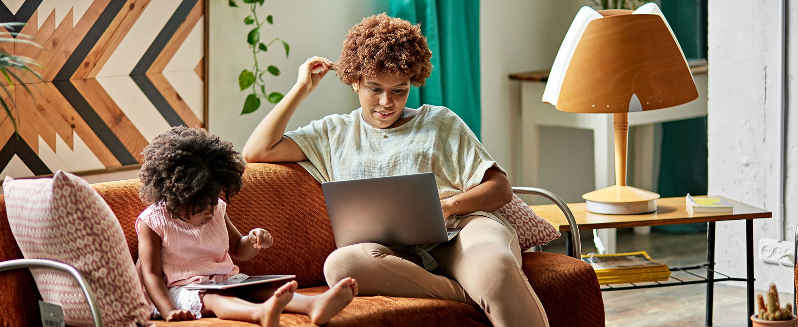 A woman works on her laptop while her daughter sits nearby on the sofa, playing with a tablet.