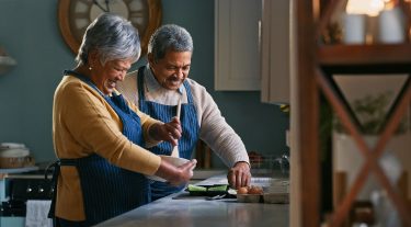 A retired couple smiles while cooking together in their kitchen.