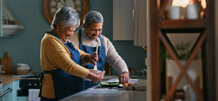 A retired couple smiles while cooking together in their kitchen.