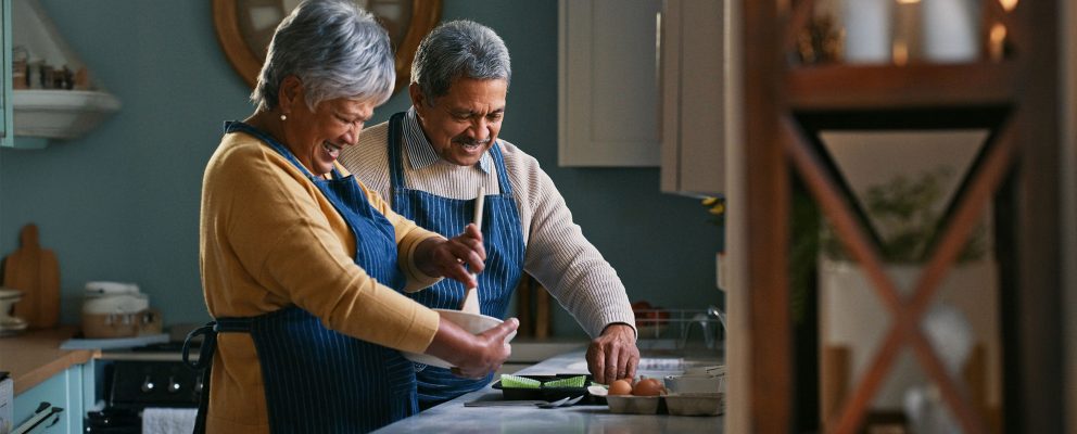 A retired couple smiles while cooking together in their kitchen.