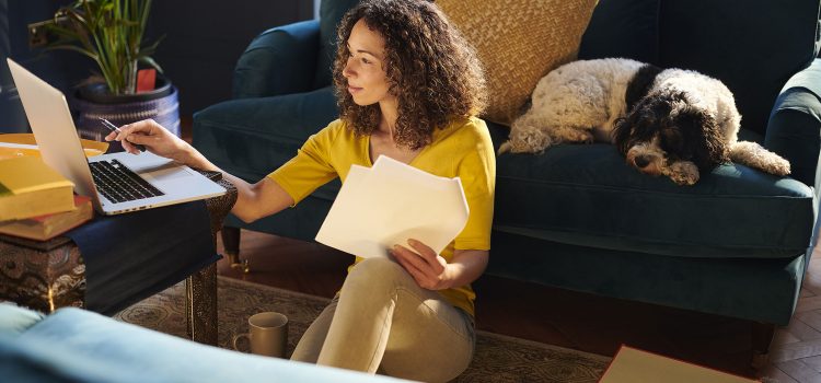 A woman works on financial paperwork at her laptop while her dog sleeps on the sofa behind her.