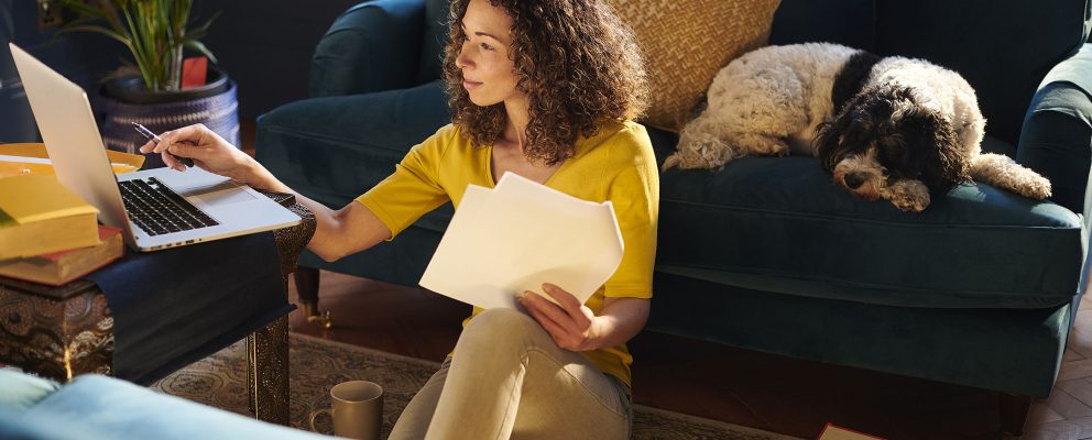 A woman works on financial paperwork at her laptop while her dog sleeps on the sofa behind her.