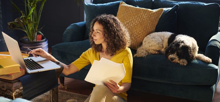 A woman works on financial paperwork at her laptop while her dog sleeps on the sofa behind her.