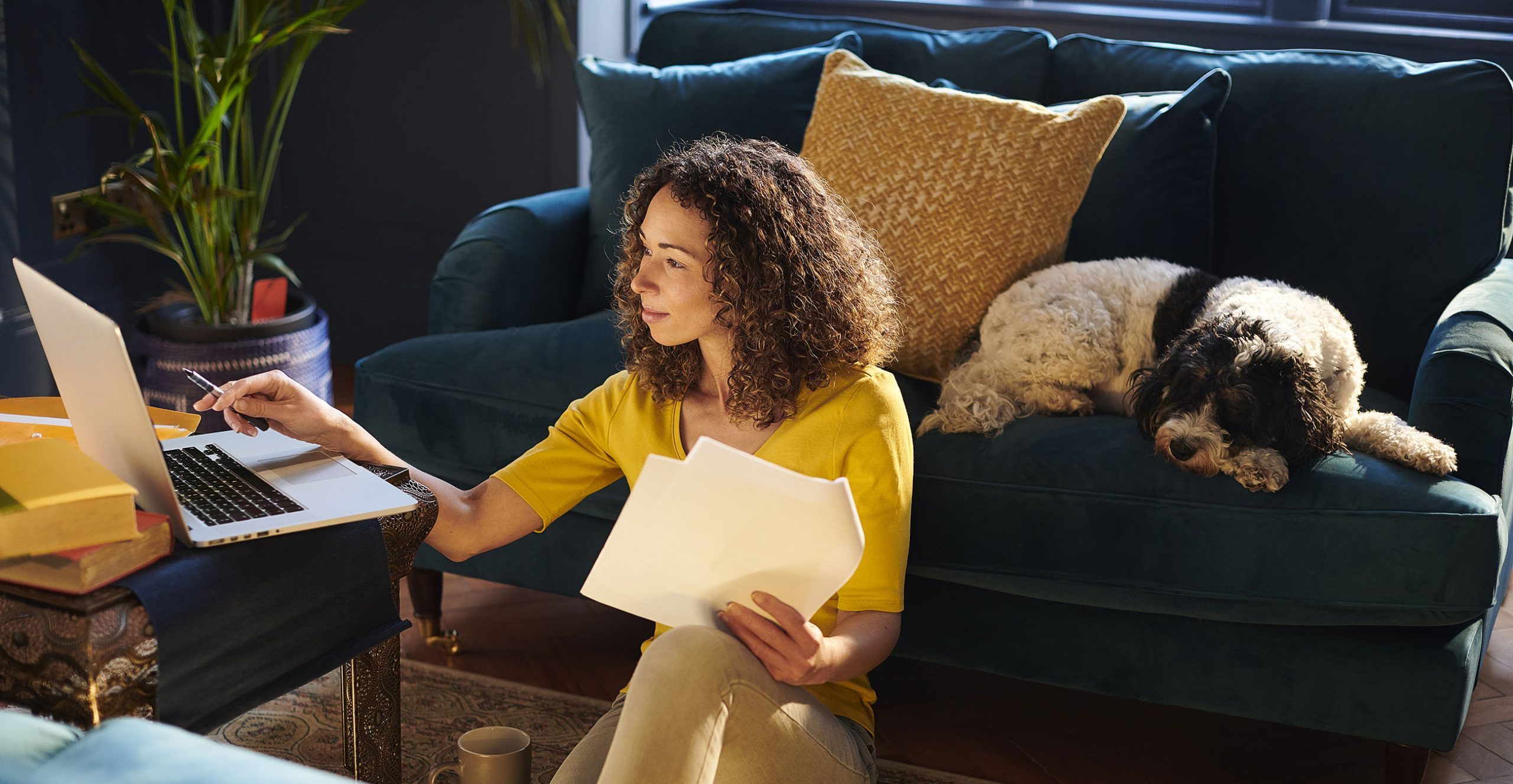 A woman works on financial paperwork at her laptop while her dog sleeps on the sofa behind her.