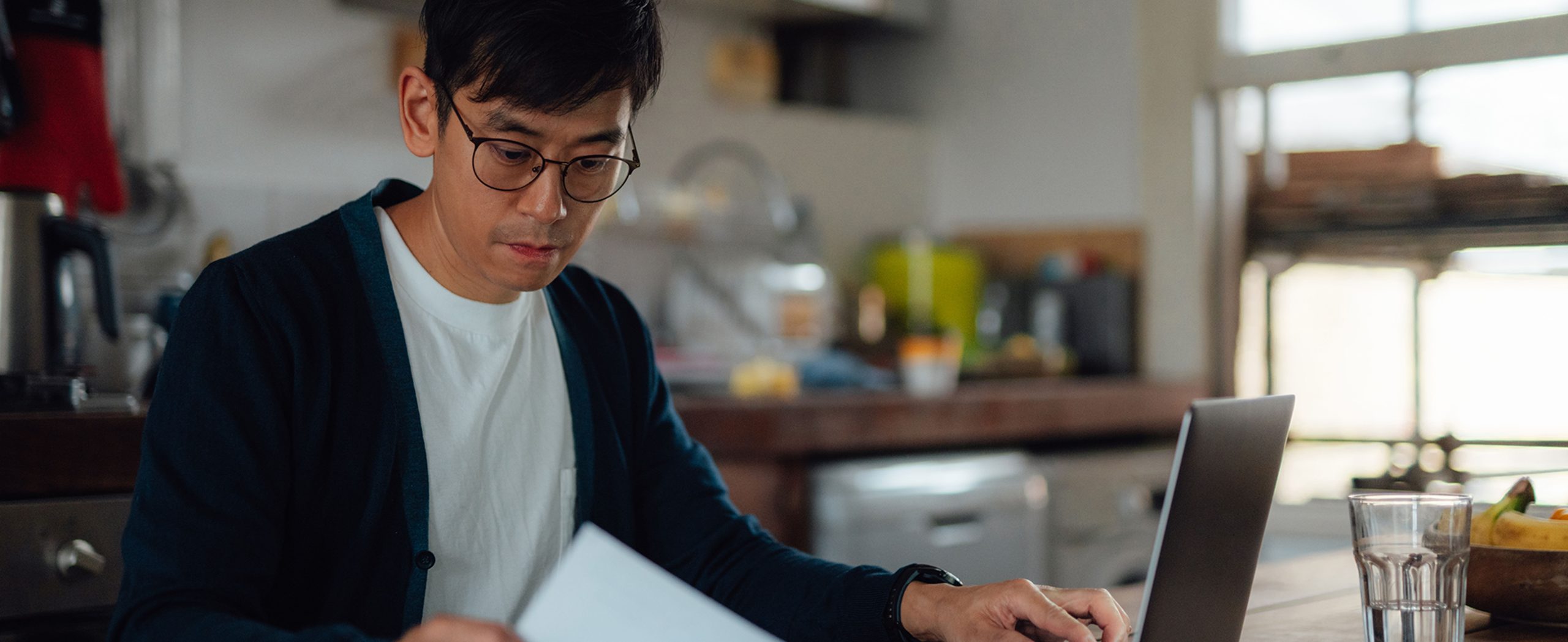 A man reviews financial paperwork next to an open laptop in his kitchen.