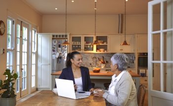 A woman discusses finances with an advisor at her kitchen table, with documents and a laptop in front of them.