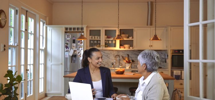 A woman discusses finances with an advisor at her kitchen table, with documents and a laptop in front of them.