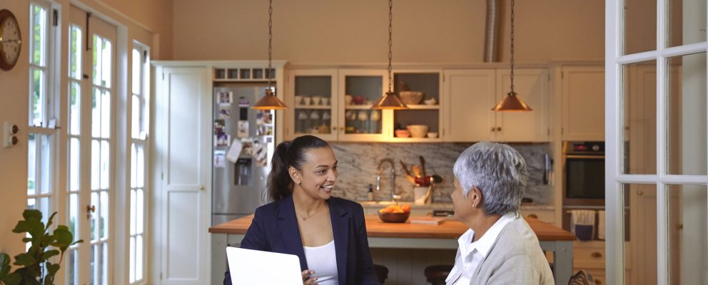 A woman discusses finances with an advisor at her kitchen table, with documents and a laptop in front of them.