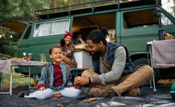 A father and son laugh outside their RV while camping while their family prepares smores in the background.
