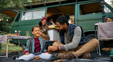 A father and son laugh outside their RV while camping while their family prepares smores in the background.