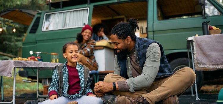 A father and son laugh outside their RV while camping while their family prepares smores in the background.