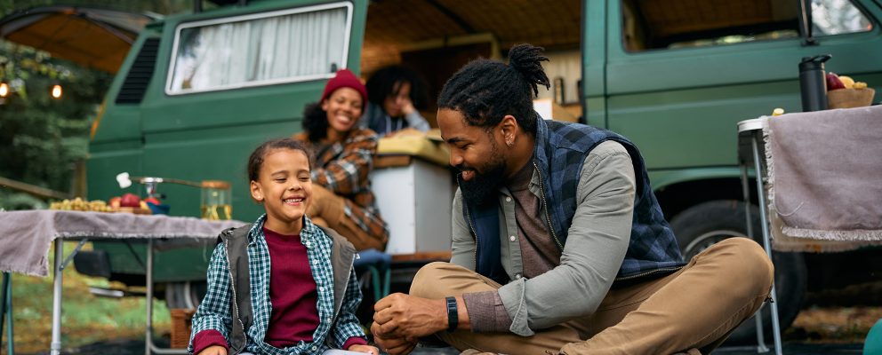 A father and son laugh outside their RV while camping while their family prepares smores in the background.