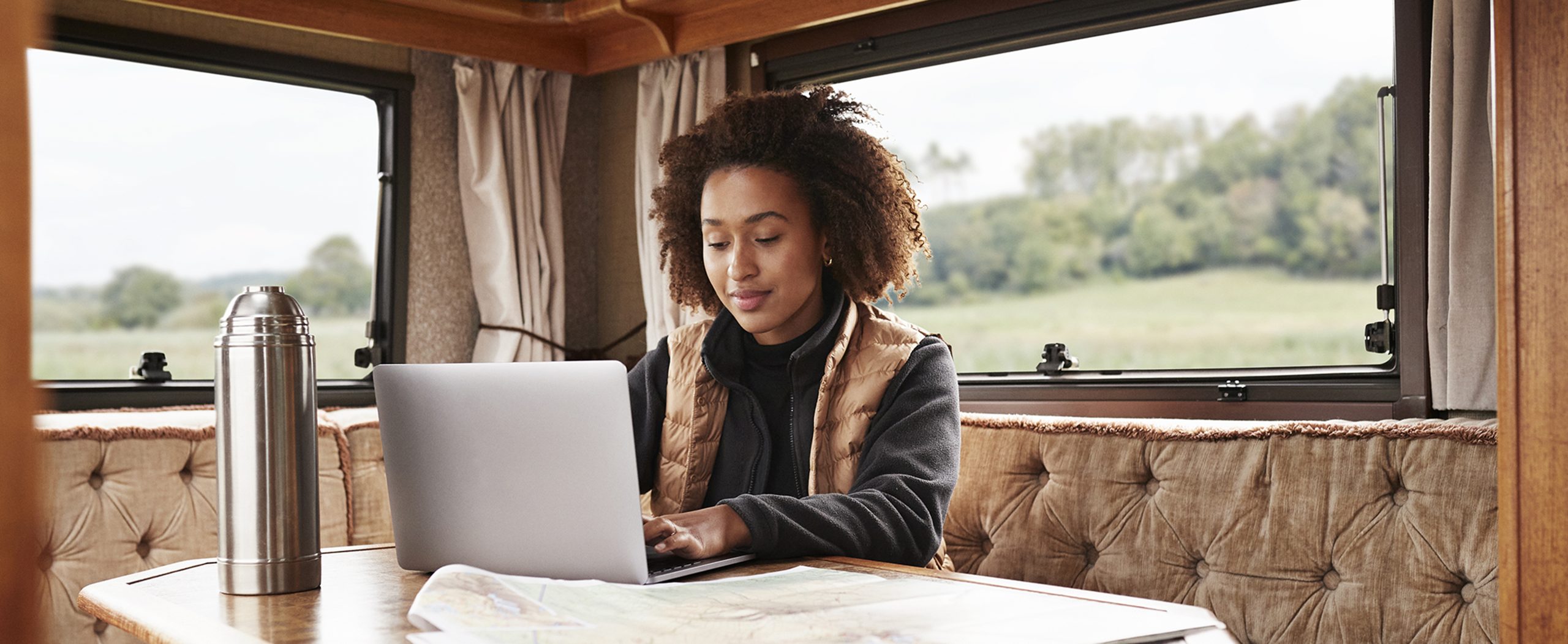 A young woman works on a laptop at a small table in her cozy camper, with a thermos of coffee beside her. 