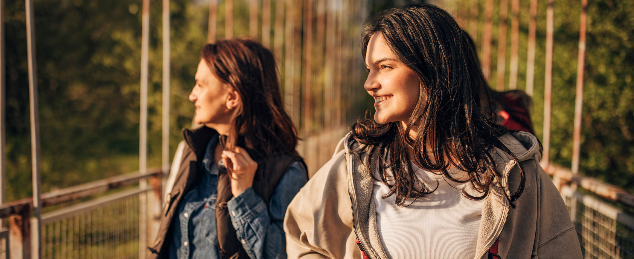 A mother and daughter walk across a bridge while hiking and look into the sun. 