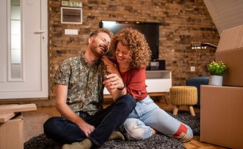 A young couple sits on the floor of their new home, laughing, next to moving boxes.