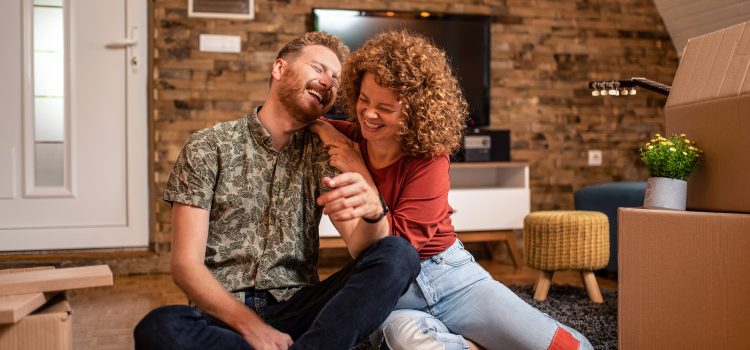 A young couple sits on the floor of their new home, laughing, next to moving boxes.