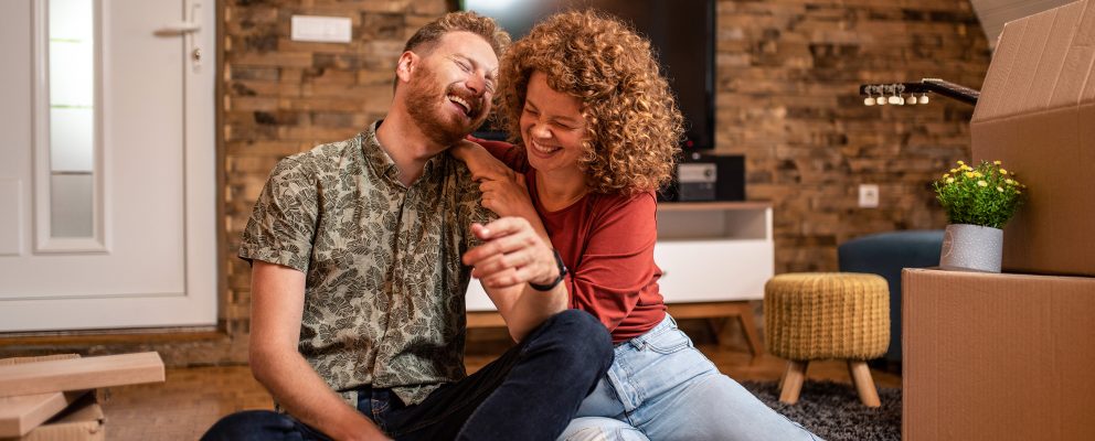 A young couple sits on the floor of their new home, laughing, next to moving boxes.