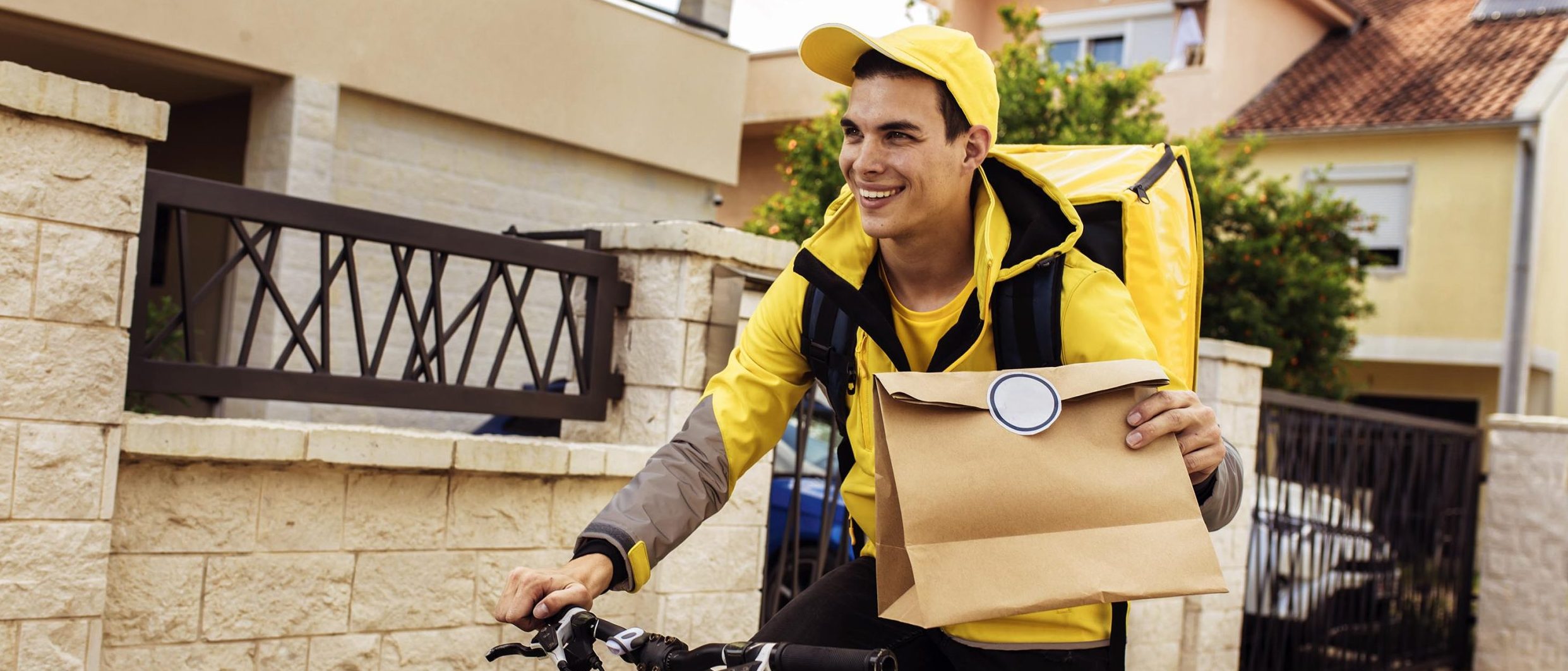 A young man smiles while riding a bike and carrying a paper bag for delivery.