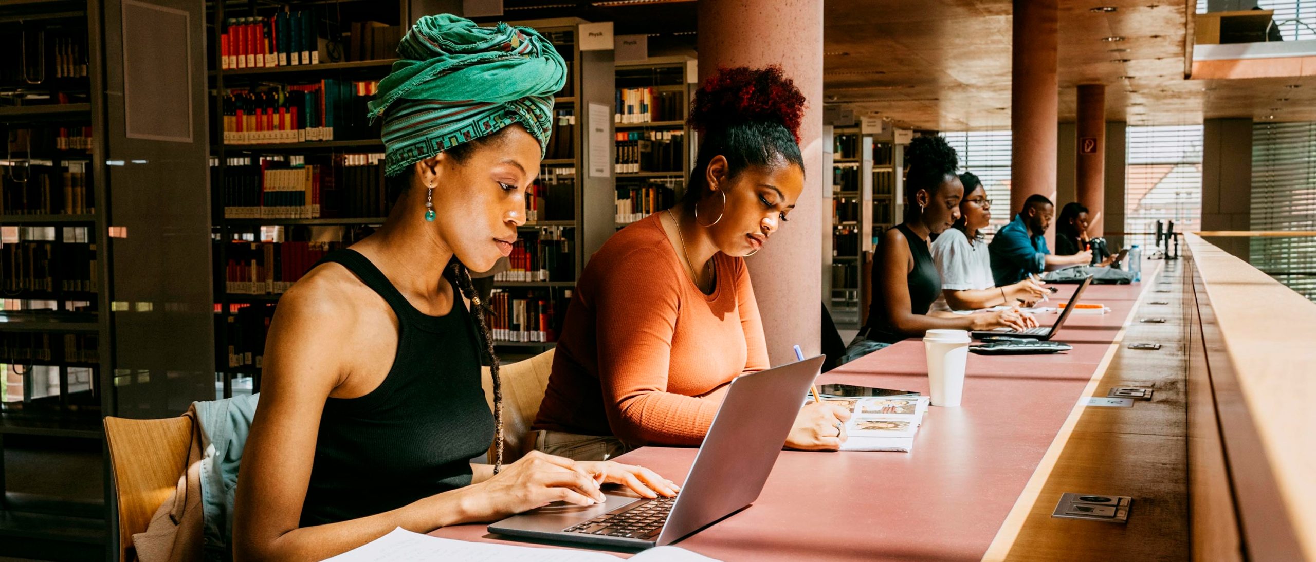 In a college library, students sit at a long table with books and laptop computers.