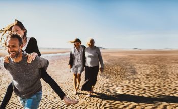 A man carries a woman on piggyback while walking on the beach, while two women walk behind with their arms around each other.
