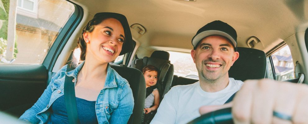 A couple smiles from the front seat of their car as their baby sits in a cars eat in the back.