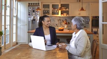 A woman discusses finances with an advisor at her kitchen table, with documents and a laptop in front of them.