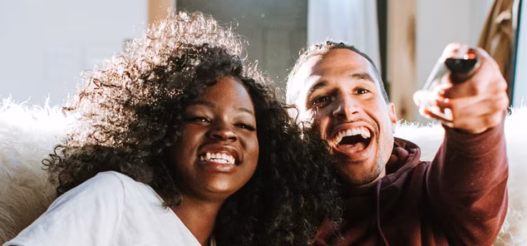 A couple sits together on the couch, laughing while watching TV.
