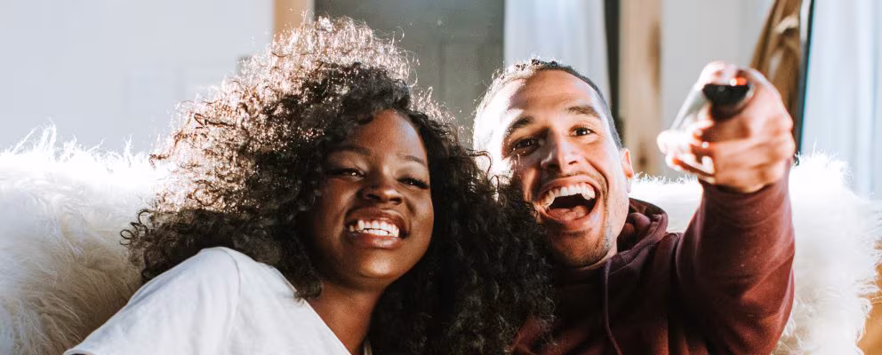 A couple sits together on the couch, laughing while watching TV.