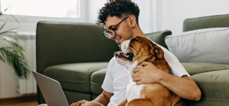 A young man sits on the floor with his laptop, hugging his dog and smiling.