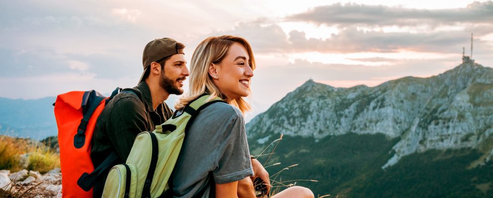 A man and woman are sitting, wearing backpacks and look out over a mountainous landscape
