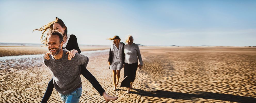 A man carries a woman on piggyback while walking on the beach, while two women walk behind with their arms around each other.