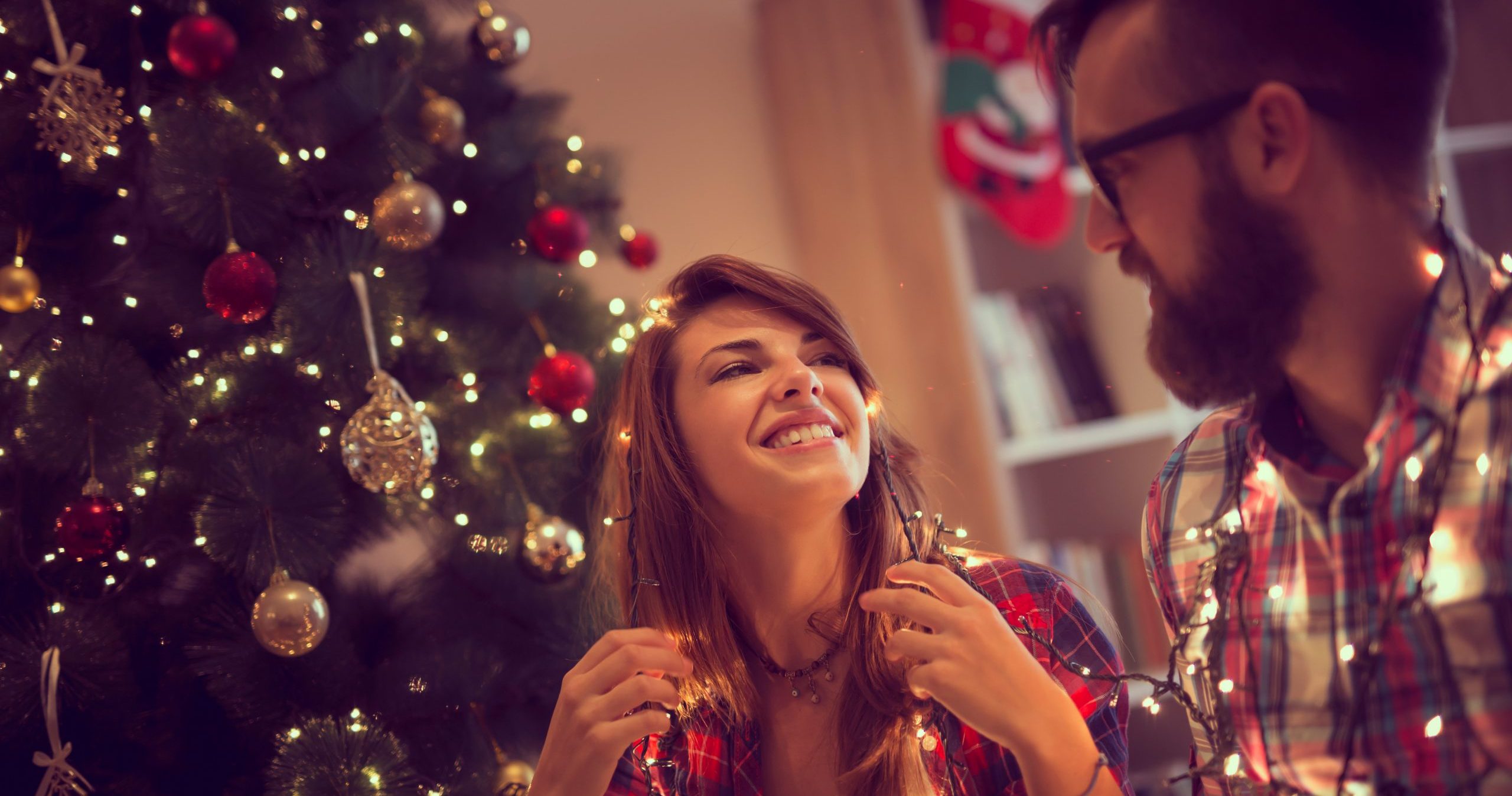 A man and a woman smile at each other while decorating a Christmas tree with a string of lights.