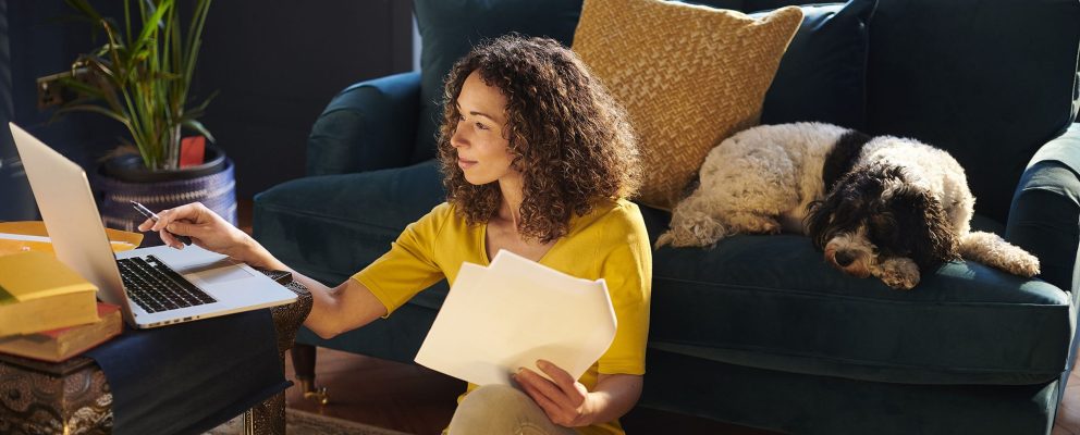 A woman works on financial paperwork at her laptop while her dog sleeps on the sofa behind her.