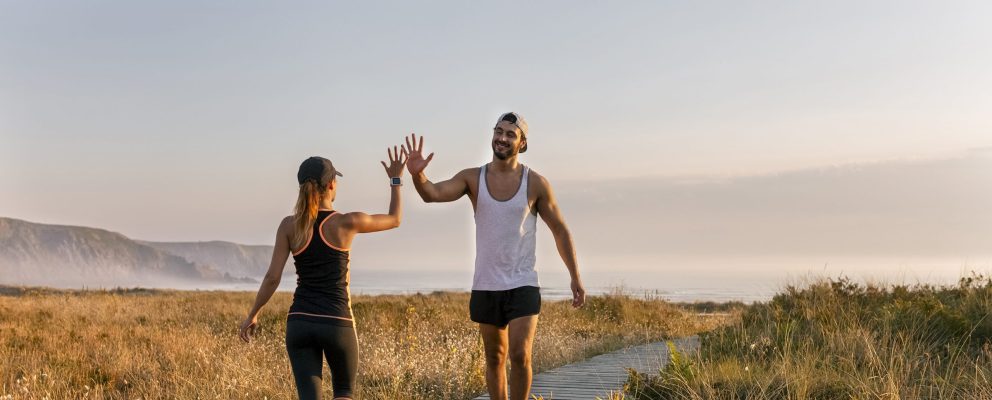A man and a woman in workout gear high five in a field by the ocean.