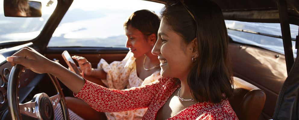 Woman driving a car while another woman sits in the passenger seat on her phone.